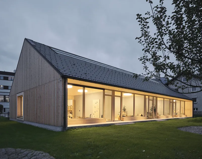 Corner view of the pitched-roof volume with illuminated glazed facade at dusk