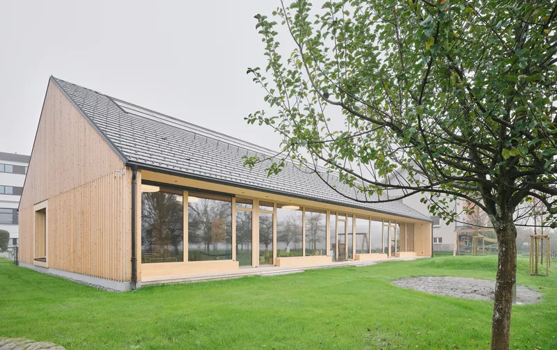 Timber-clad gable building with full-height glazed facade framed by a deciduous tree on a lawn