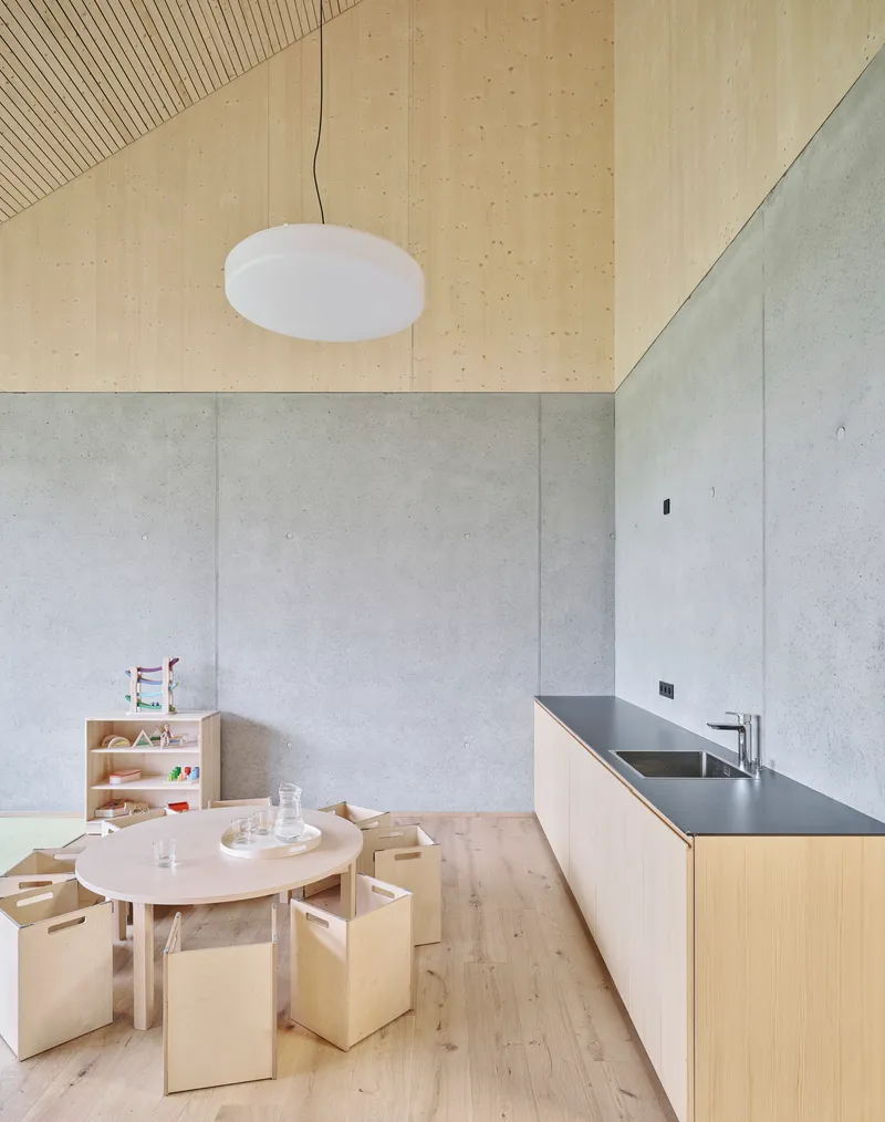 Interior view of children's space with pale concrete walls, plywood ceiling and kitchenette with dark countertop