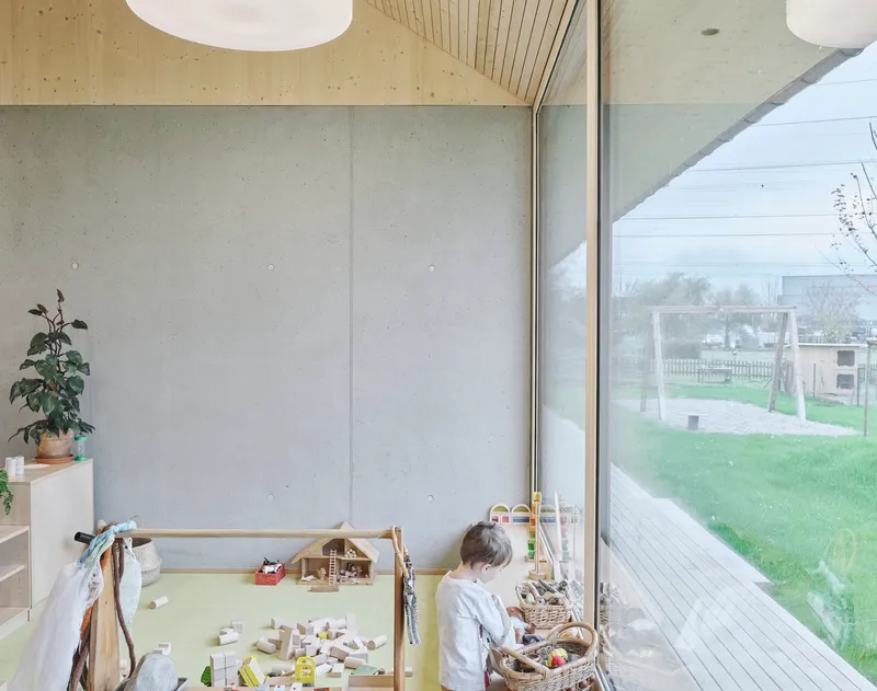 Child playing at wooden play table beside full-height glazing overlooking garden and playground equipment