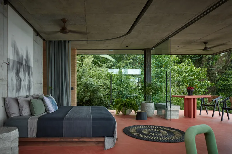 Bedroom interior with concrete ceiling opening to red terrace and lush garden through sliding glass