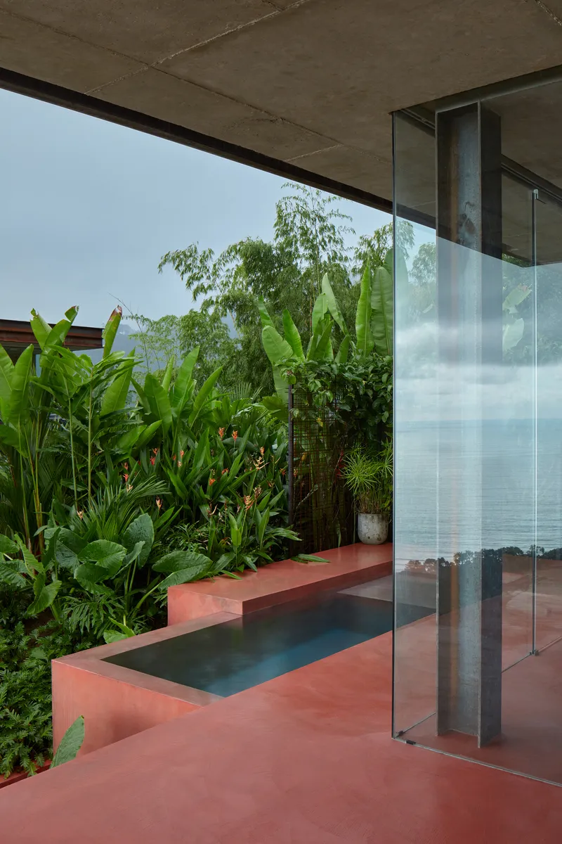 Covered terrace with red concrete flooring, reflecting pool, and dense tropical plantings beyond glass