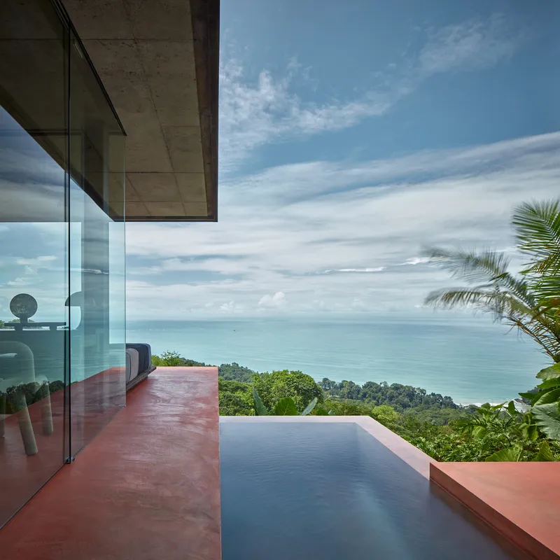 Red concrete terrace with infinity pool facing ocean and forested coastline under scattered clouds