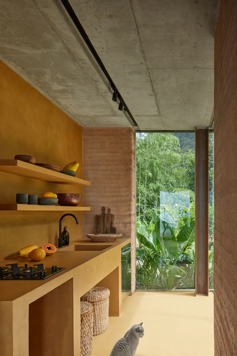 Kitchen interior with ochre plaster walls and concrete ceiling framing a view of lush tropical vegetation