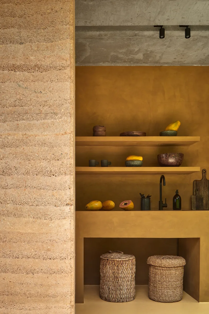 Kitchen alcove with floating timber shelves against ochre plaster walls beside a rammed earth partition