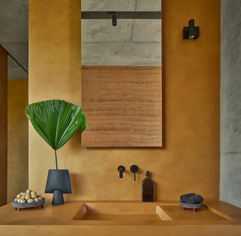 Bathroom sink detail with black tapware and framed rammed earth panel above ochre plaster countertop