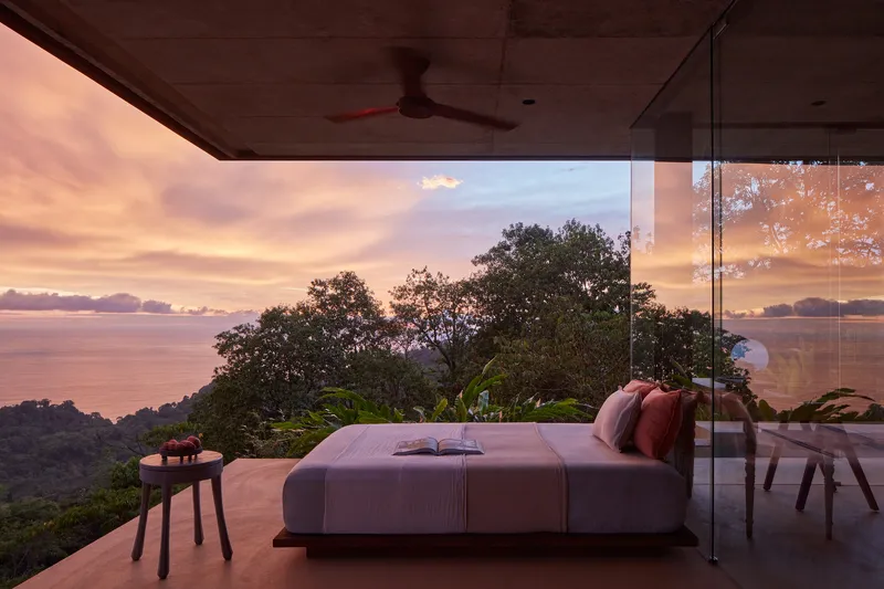 Open-air bedroom with glass walls framing treetops and ocean at sunset