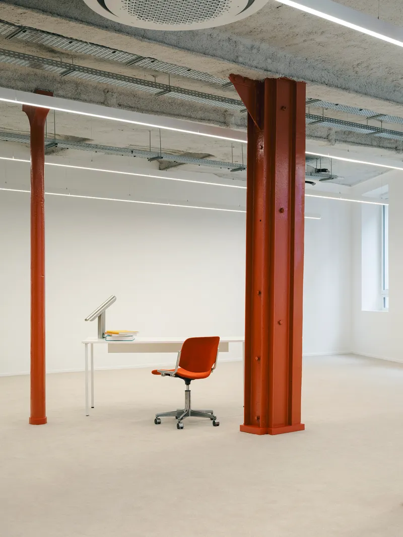 Orange steel column beside minimal white desk and chair beneath exposed concrete ceiling with linear lighting