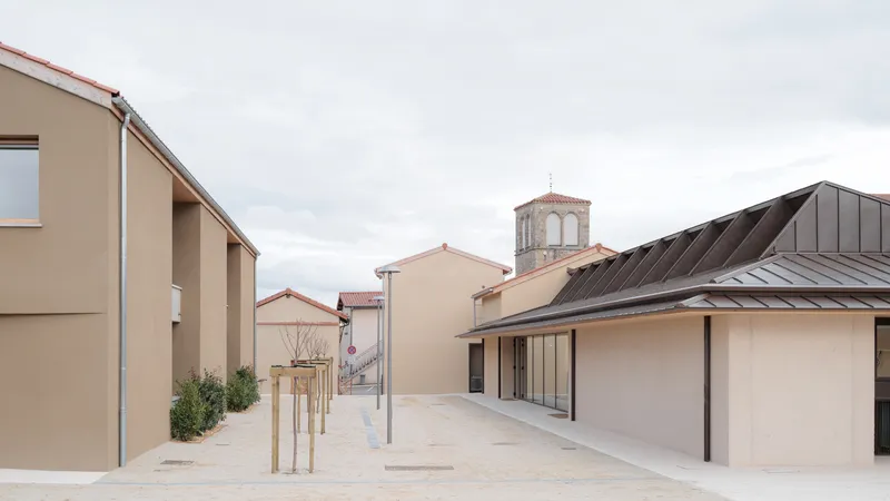 Open courtyard flanked by beige stucco volumes with a row of young trees and a church tower beyond