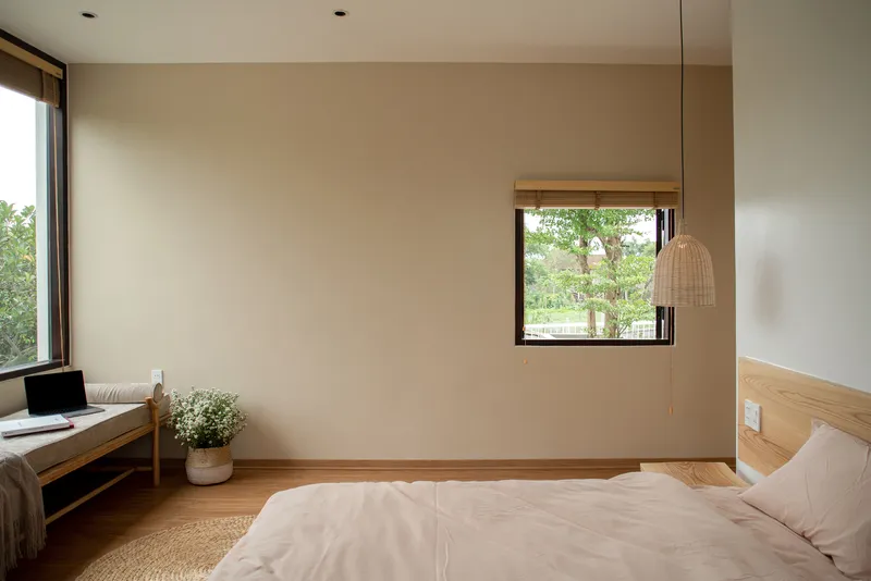 Bedroom with pale walls, timber bed frame, woven pendant light, and window overlooking garden foliage