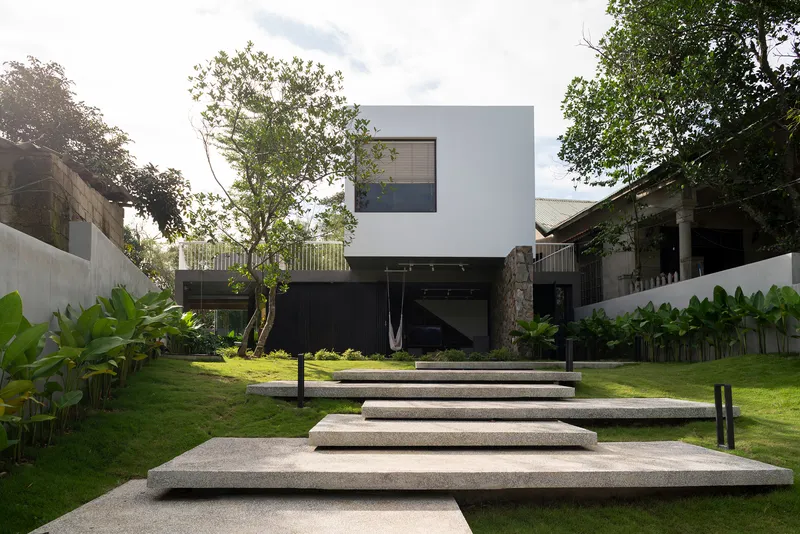 Terrazzo platform steps intersecting the lawn with white cubic upper volume framed by mature trees overhead