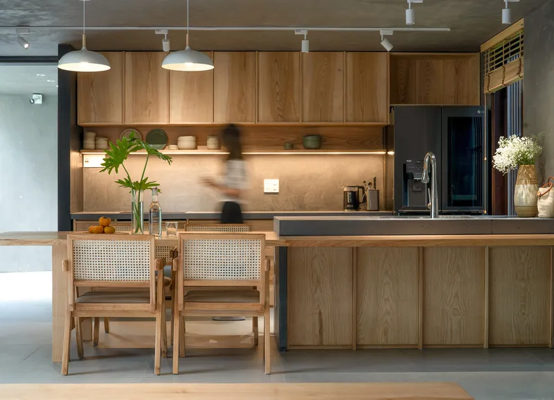 Kitchen island with cane-backed chairs beneath timber cabinetry and track lighting on exposed concrete ceiling