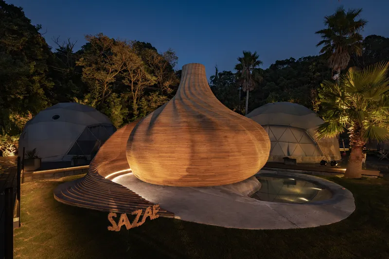 Teardrop-shaped timber pavilion beside two geodesic domes and palm trees at twilight