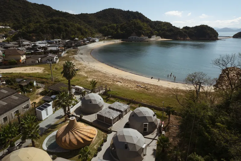 Cluster of geodesic domes and ribbed timber pavilion overlooking a coastal bay with forested hills