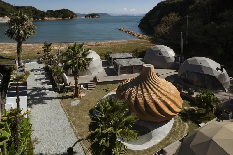 Aerial view of cluster of geodesic domes and ribbed timber shell structure on coastal lawn with palm trees