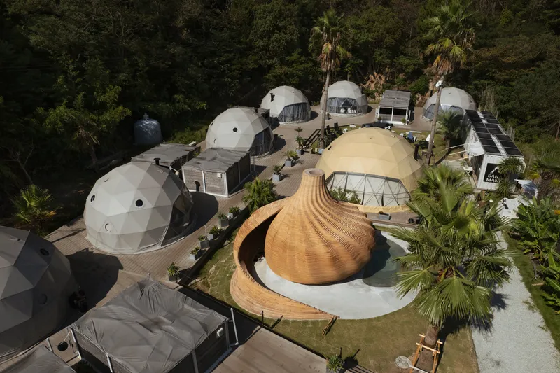 Overhead view of bulbous timber shell pavilion surrounded by white geodesic domes and rectangular modules among palms