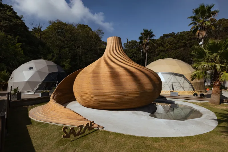 Ribbed timber shell pavilion with chimney-like opening and curving base surrounding a reflecting pool in bright sun