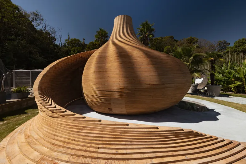 Laminated timber ribs forming bulbous pavilion with extended canopy deck on white concrete base under blue sky