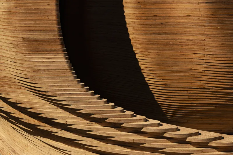 Close-up of curved laminated timber ribs casting sharp shadow across horizontal wood slat surface in afternoon light