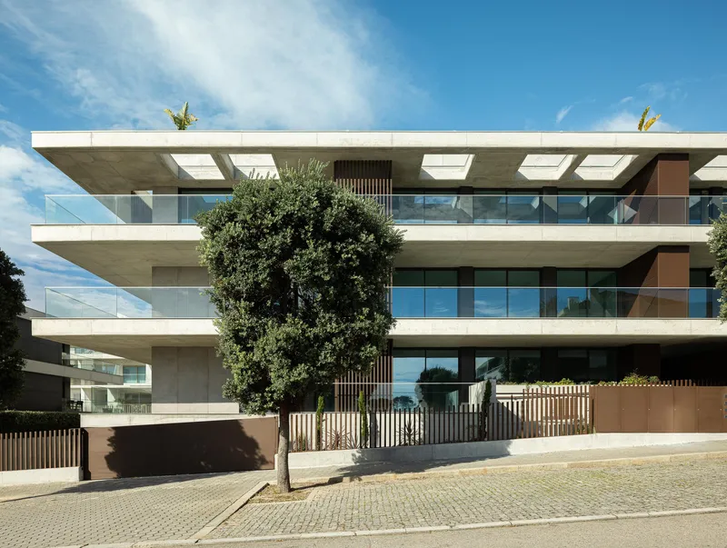 Street facade with cantilevered concrete balconies and glass railings framed by mature trees under blue sky