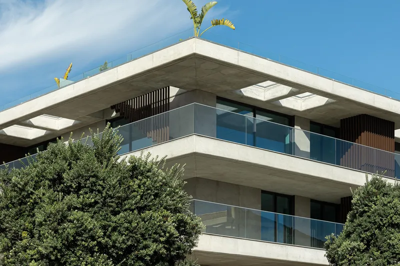Corner detail of concrete balconies with glass railings, vertical timber screens, and rooftop banana plants