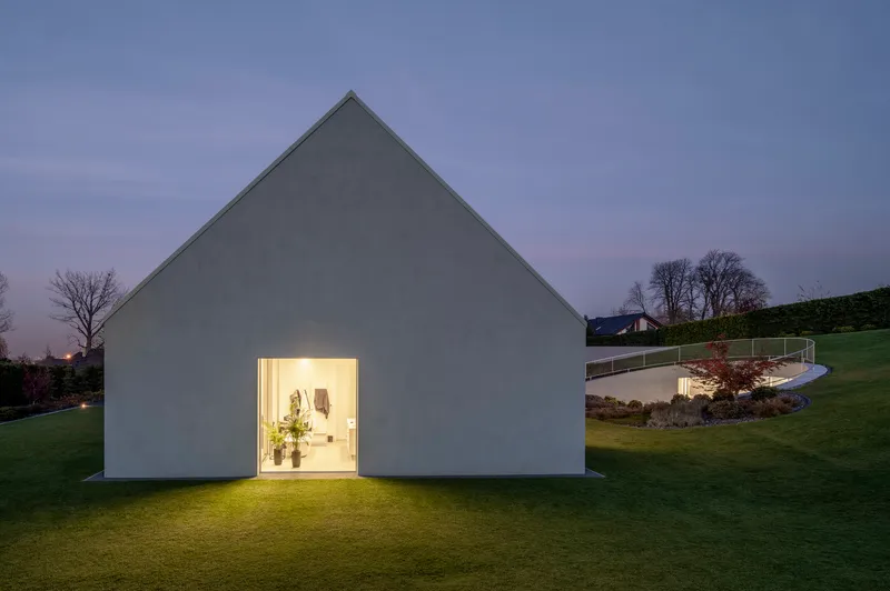 Gabled white volume with single illuminated doorway opening onto lawn at dusk