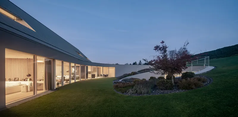 Glass wall revealing interior living space with curving planted bed at twilight