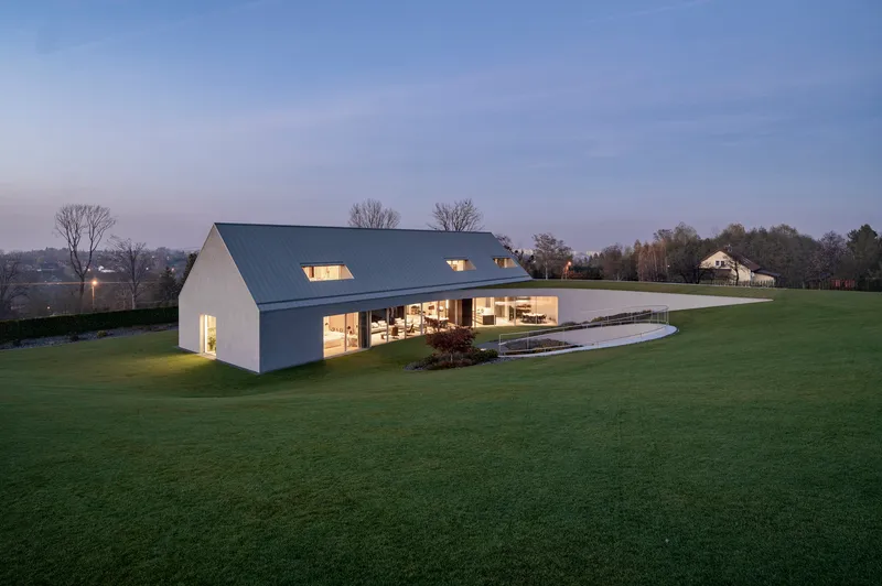 Gabled house with metal roof and glazed lower level set into sloping lawn at dusk