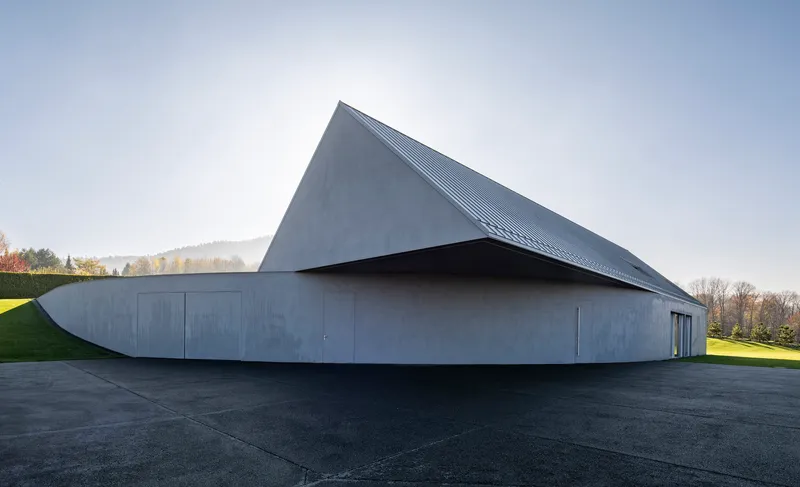 Angular zinc roof volume cantilevering over curved concrete base with flush doors facing the lawn