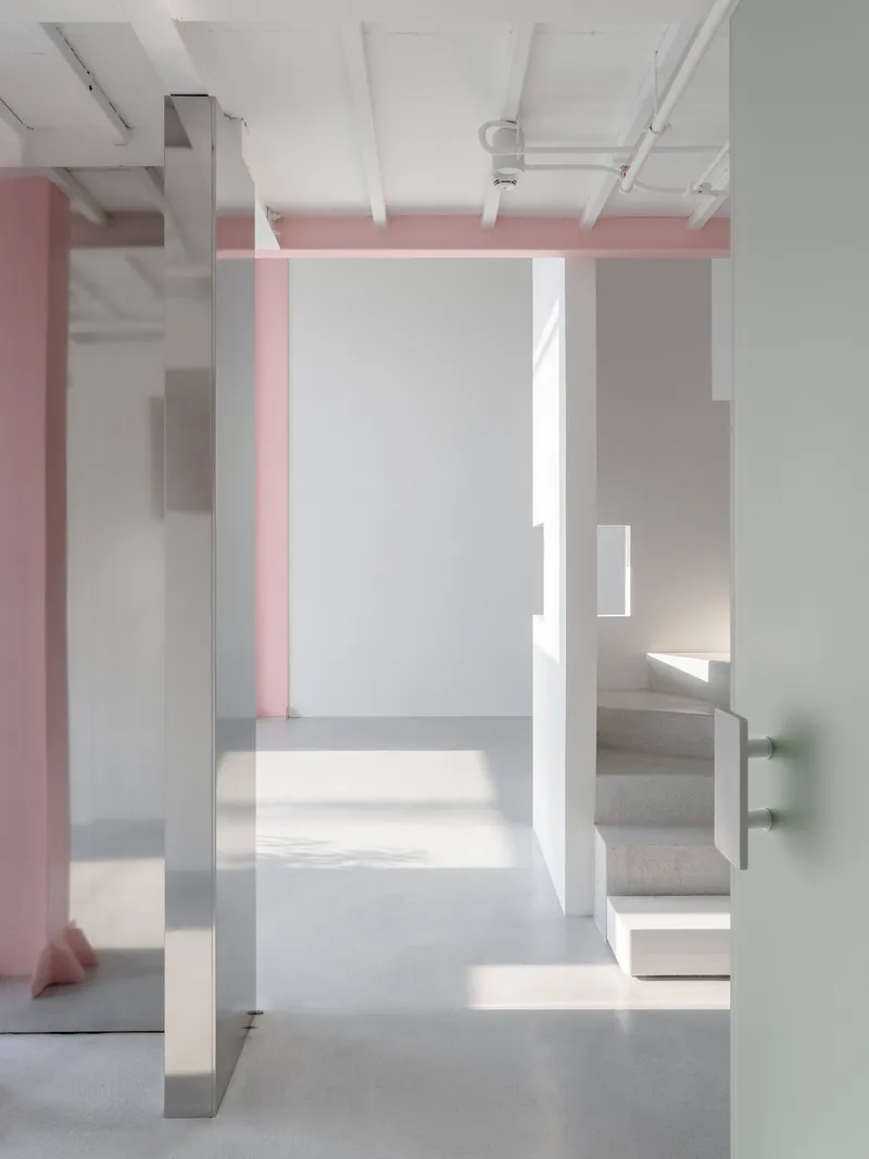 Interior view through white partitions with pink beams overhead and sunlight casting geometric shadows on polished concrete floors