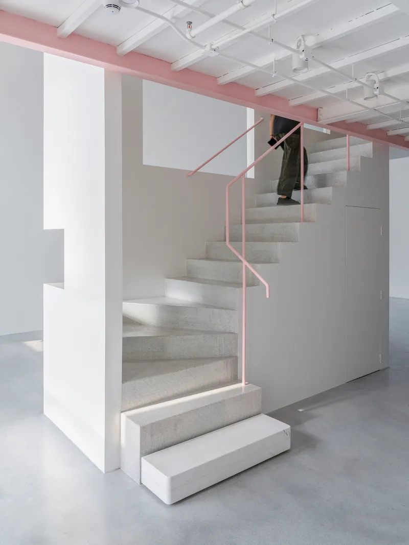 Carpeted staircase with pink tubular steel handrail ascending beneath exposed white coffered ceiling with pink structural beam