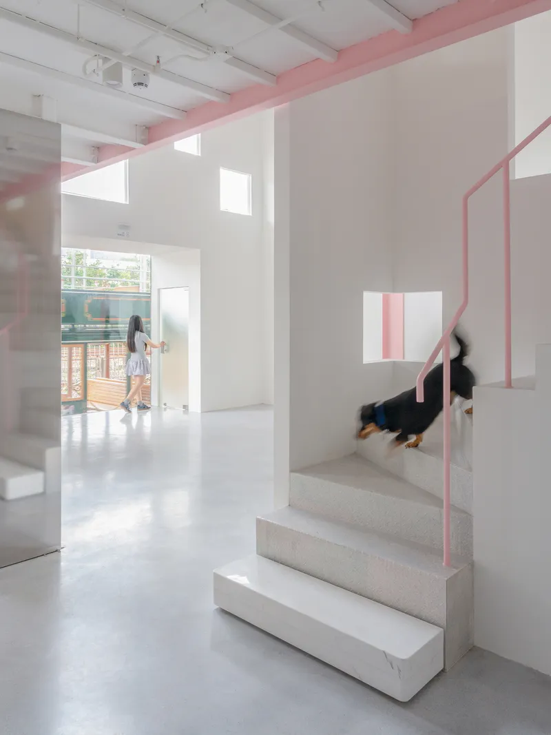Open stairwell with person descending pink-railed stairs while another figure walks toward windowed entrance in bright daylight