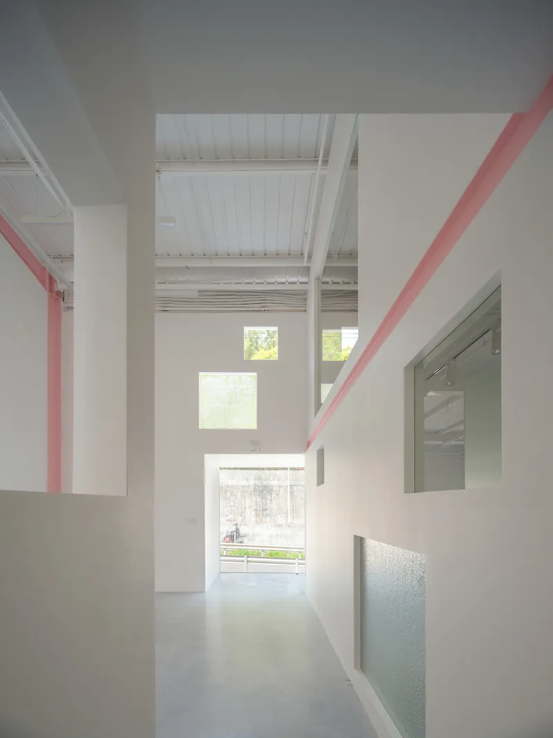 Corridor view through white partition walls with pink diagonal beams and stacked windows framing green foliage outside