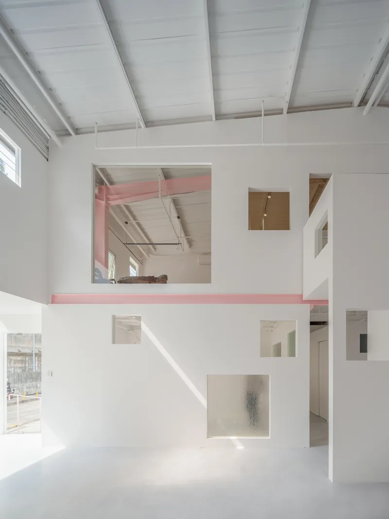 Double-height interior with white partition walls punctured by square openings and pink beam spanning upper mezzanine level