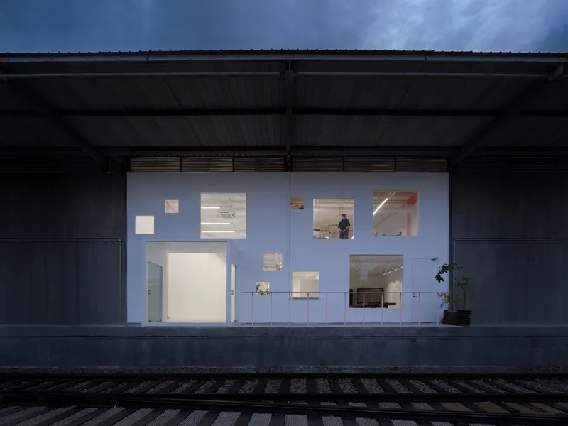 Illuminated white facade wall beneath a metal shed canopy at dusk with a figure visible inside