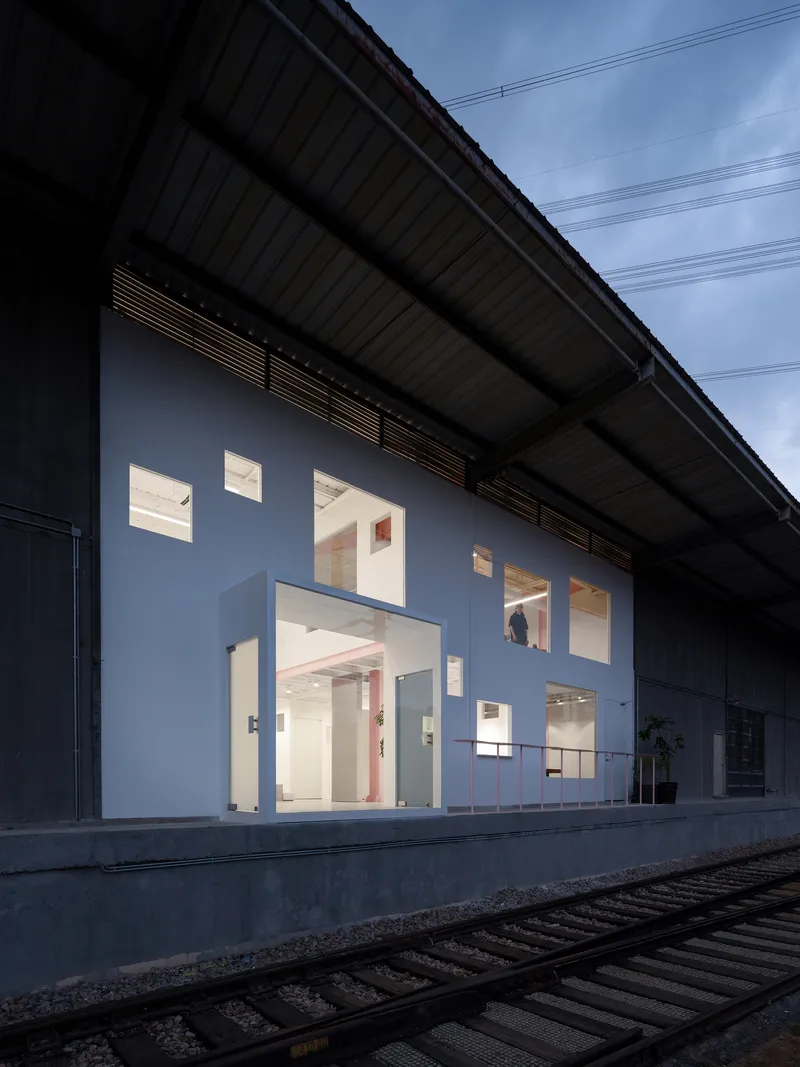 White facade wall with scattered window openings under a corrugated metal canopy and railway tracks in foreground