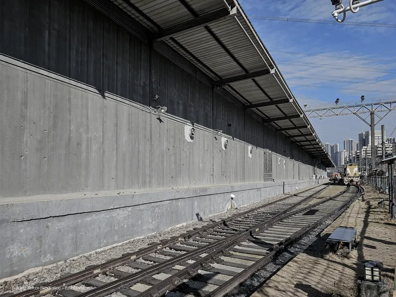 Long concrete wall with metal canopy running parallel to railway tracks under clear blue sky