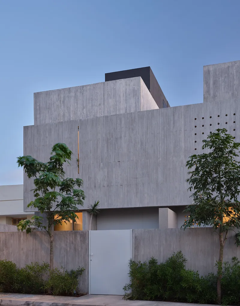 Stacked concrete volumes with perforated panel openings and a white entry gate flanked by young trees at dusk