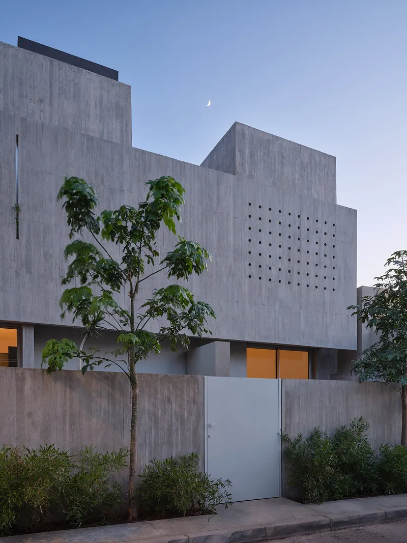 Board-formed concrete facade with perforated wall panel and a crescent moon visible above the stepped volumes at twilight