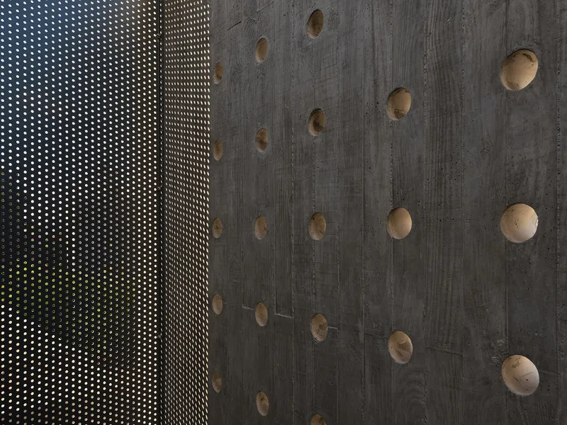 Perforated metal screen beside board-formed concrete wall with exposed tie holes and green foliage beyond