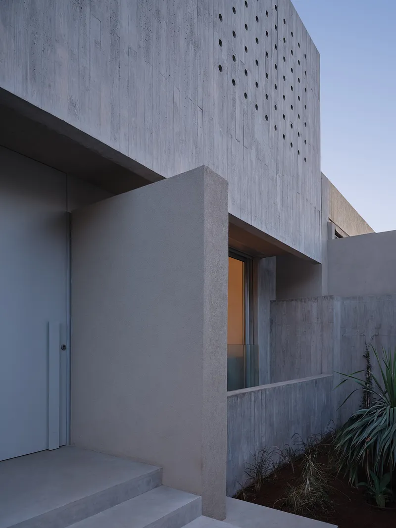 Concrete facade with scattered circular perforations at dusk beside planted beds with spiky grasses