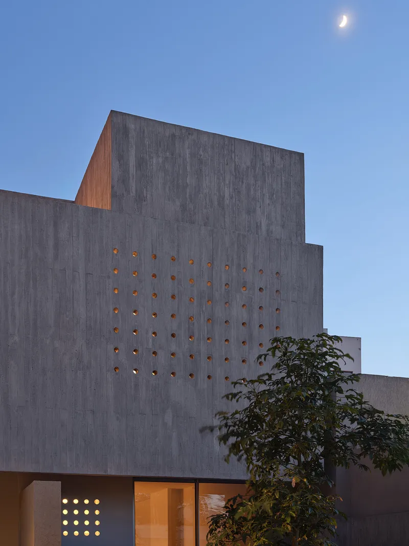Stepped concrete facade with perforated openings at dusk with crescent moon above and tree beside entrance
