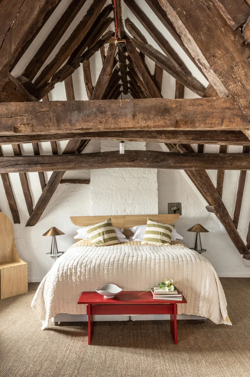 Bedroom beneath exposed timber roof trusses with a red bench at the foot of the bed