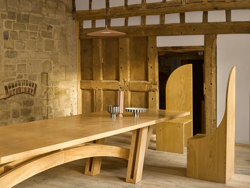 Oak dining table with curved plywood chairs beneath exposed timber frame and stone walls
