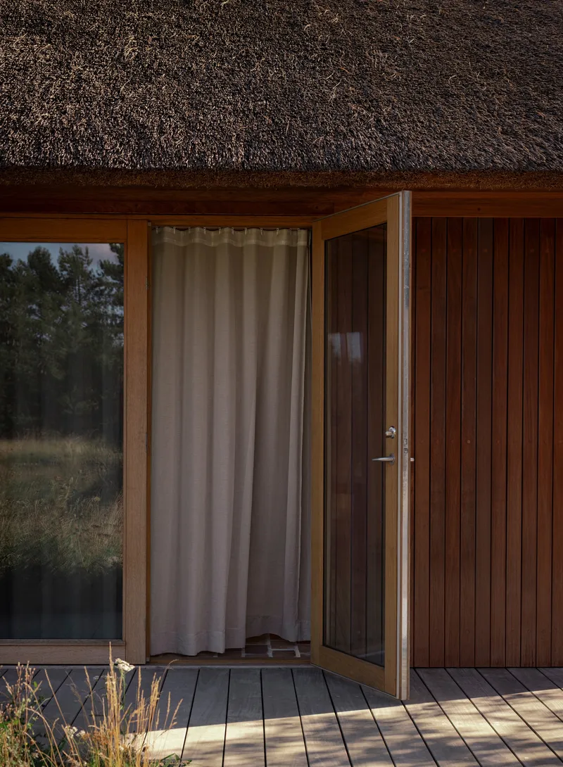 Timber deck threshold with open glass door and linen curtain under the thatched eave