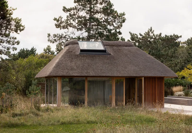 Thatched roof pavilion with timber cladding and full-height glazing surrounded by wild grasses and pine trees
