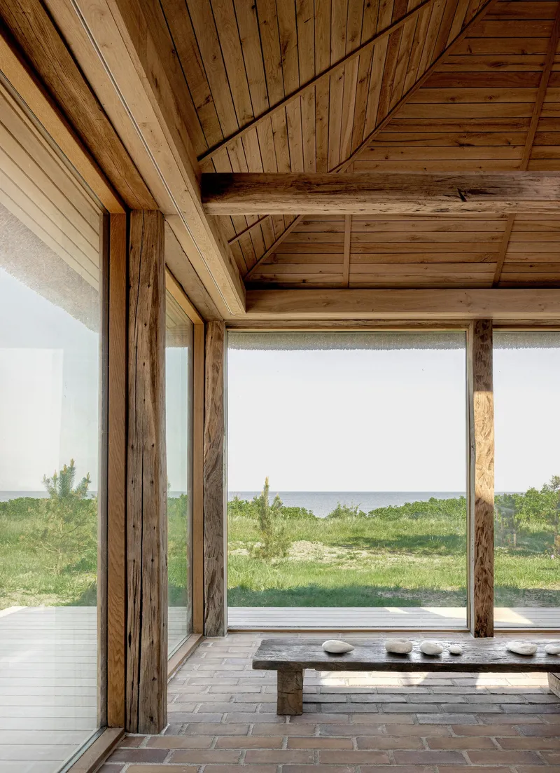 Interior corner with timber columns, planked ceiling and full-height glazing overlooking coastal dunes