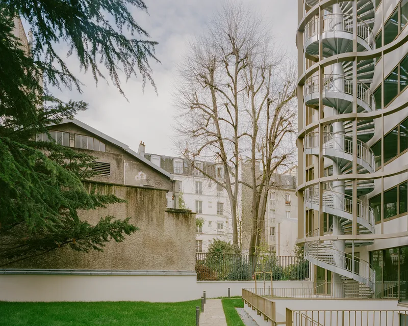 Curved facade with exposed spiral staircases beside a bare deciduous tree in a courtyard lawn