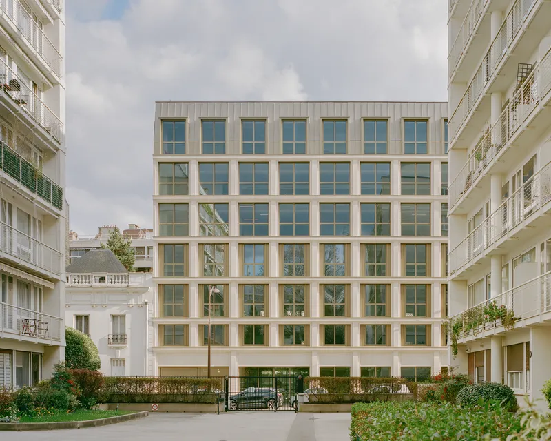 Grid facade with beige stone frames and paired windows viewed from a landscaped courtyard under overcast skies