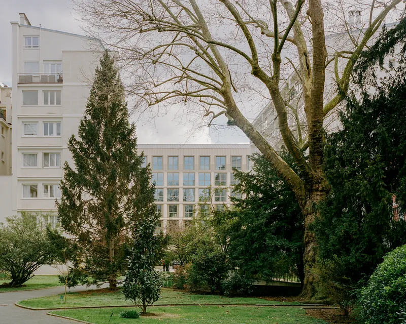 Garden view of the gridded facade framed by evergreen and deciduous trees on an overcast day